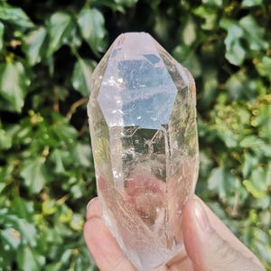 May include: A clear quartz crystal point with a natural, faceted shape. The crystal is translucent and has a slight pink hue. It is approximately 10 cm tall.