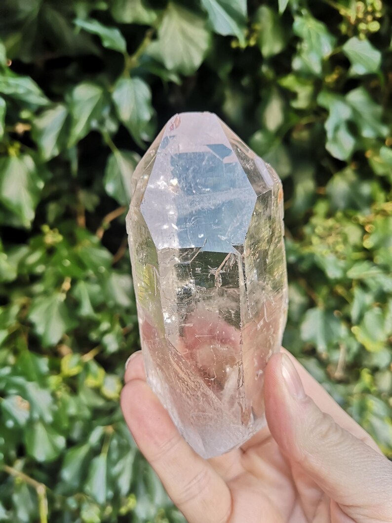 May include: A clear quartz crystal point with a natural, faceted shape. The crystal is translucent and has a slight pink hue. It is approximately 10 cm tall.