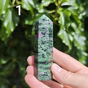 May include: A polished hexagonal crystal tower featuring a mottled green and black base with embedded vibrant magenta-pink inclusions. The stone has a smooth, faceted surface that tapers to a point at the top, marked with the number 1.