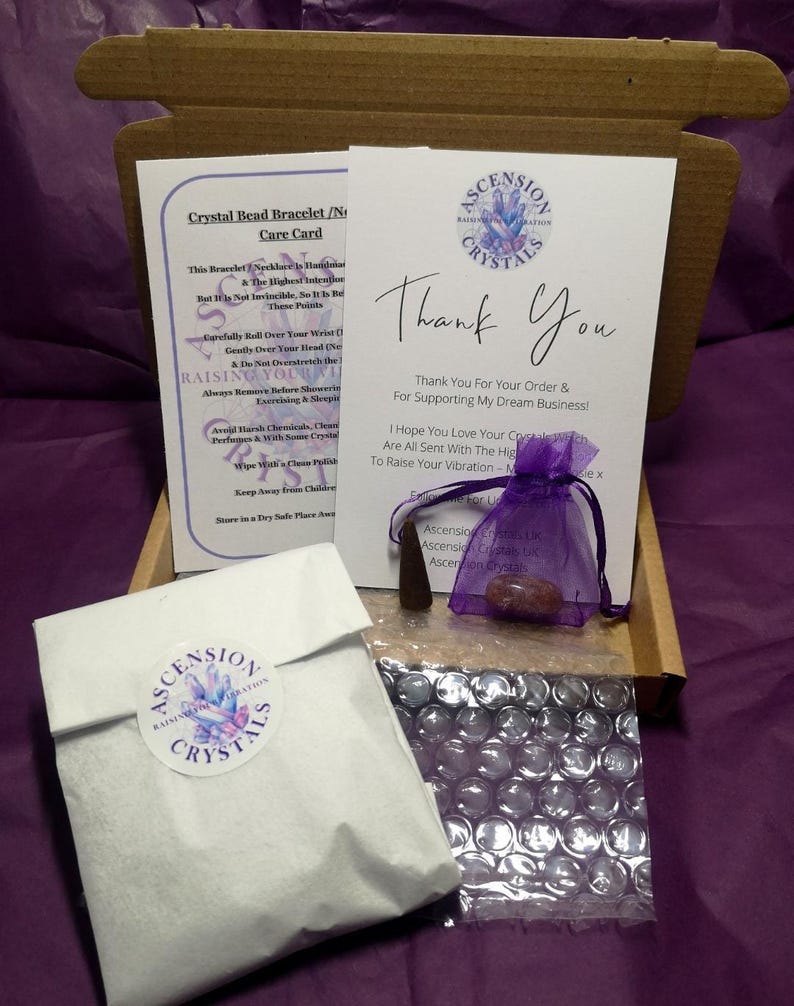 May include: A cardboard shipping box containing a white care card, a white thank you card with the Ascension Crystals logo, a small purple organza drawstring bag with a crystal, a dark cone-shaped incense, and a white paper pouch with a logo sticker.