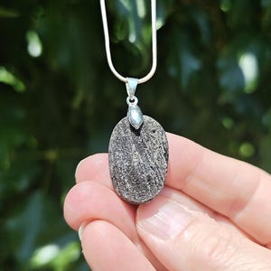 May include: An oval-shaped, dark grey, textured stone pendant suspended from a thin silver-toned chain. The stone features visible horizontal striations and a rough, granular surface, held by a polished silver-toned metal bail at the top.