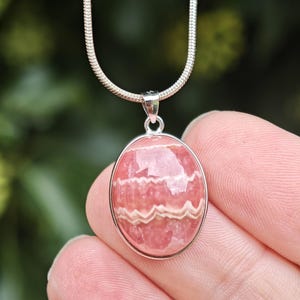 May include: An oval-shaped rhodochrosite pendant featuring distinct pink and white banded patterns, set in a polished silver-toned metal bezel. The pendant hangs from a matching silver-toned snake chain necklace with a smooth, rounded link design.