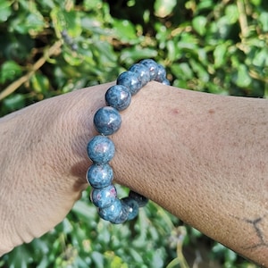 Peut inclure: Un bracelet de perles composé de pierres rondes et polies dans des tons bleu profond et gris, avec des taches occasionnelles de rouge foncé. Les perles sont enfilées en une boucle circulaire continue, présentant une texture de surface lisse.