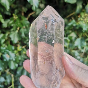 May include: A clear quartz crystal point with a natural, raw finish. The crystal is approximately 12.5 cm tall and has a hexagonal shape. The point is clear and translucent, with some inclusions visible.