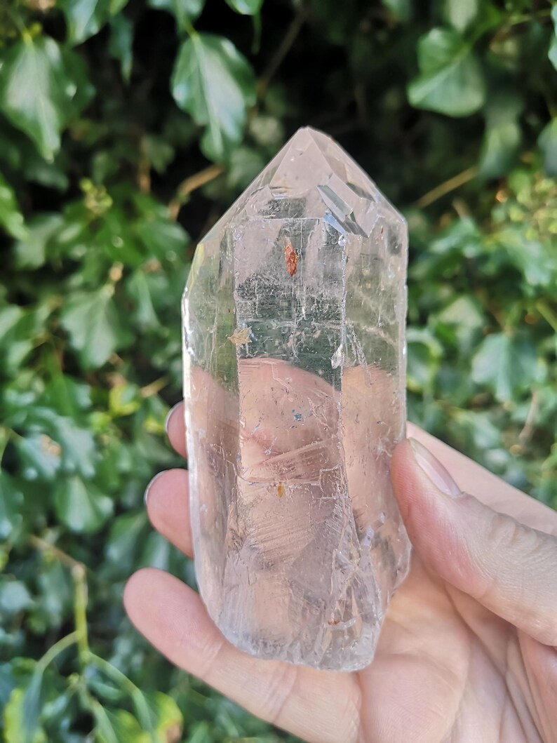 May include: A clear quartz crystal point with a natural, raw finish. The crystal is approximately 12.5 cm tall and has a hexagonal shape. The point is clear and translucent, with some inclusions visible.