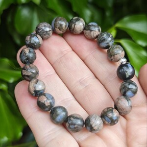 Peut inclure: Un bracelet de perles rondes et texturées, de couleurs grises, marron et bleues. Les perles sont tenues dans une main, mettant en valeur les variations de texture et de couleur de la pierre naturelle. Chaque perle mesure environ 1,3 cm de diamètre.