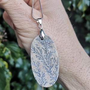 May include: A silver chain necklace with an oval pendant made of a light brown stone with a dark gray dendritic pattern.