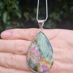 May include: A teardrop-shaped pendant necklace featuring a green, pink, and gold gemstone. The pendant is set in silver and hangs from a silver chain.