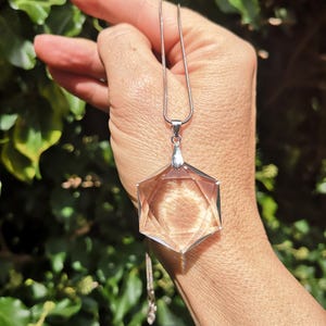 May include: A clear, faceted crystal pendant in a Star of David shape, suspended from a silver chain. The pendant is held up against a backdrop of green foliage, showcasing its geometric design and clarity.