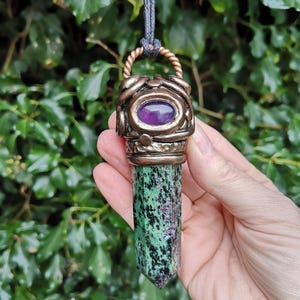 May include: A pendant necklace with a green and black zoisite crystal. The pendant has a bronze-coloured metal setting with a purple gemstone. The necklace is suspended from a grey cord.
