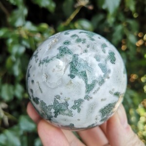 May include: A green and white moss agate sphere. The sphere is smooth and polished, with a swirling pattern of green and white.