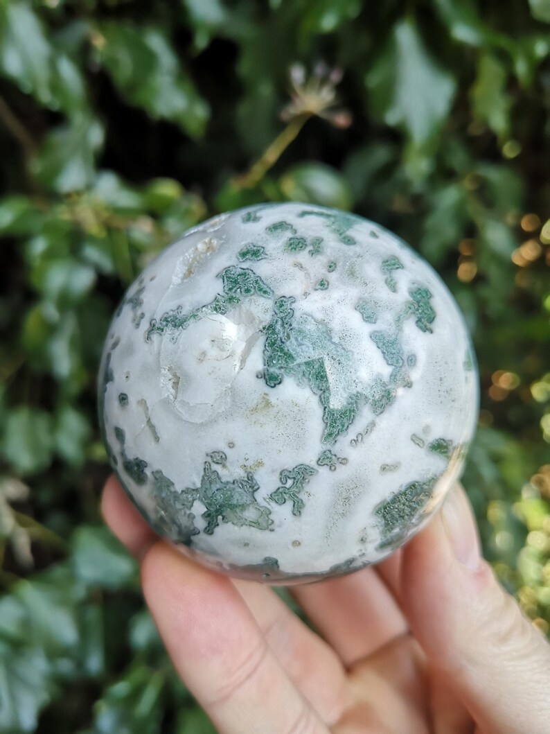 May include: A green and white moss agate sphere. The sphere is smooth and polished, with a swirling pattern of green and white.
