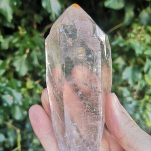 May include: A clear quartz crystal point with a natural, raw finish. The crystal is approximately 12.5 cm tall and has a hexagonal shape. The point is clear and translucent, with a slight yellow hue.