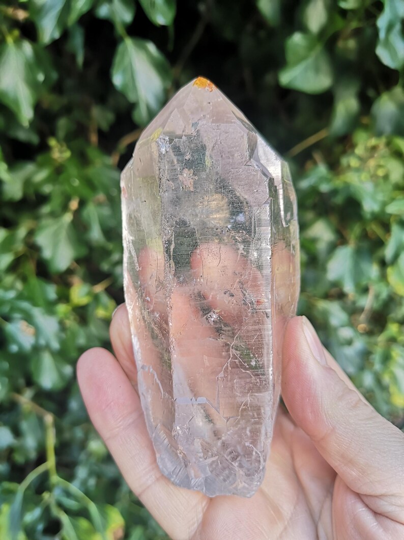 May include: A clear quartz crystal point with a natural, raw finish. The crystal is approximately 12.5 cm tall and has a hexagonal shape. The point is clear and translucent, with a slight yellow hue.