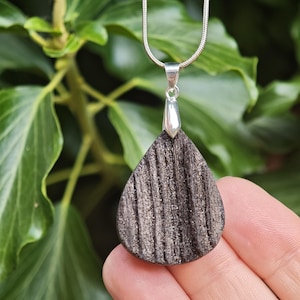 May include: A teardrop-shaped pendant featuring a dark, textured surface with vertical grooves and a metallic sheen. The pendant is attached to a silver-toned snake chain by a polished silver bail, creating a cohesive piece of jewelry.