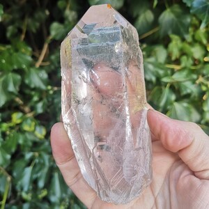 May include: A large, clear quartz crystal with a pointed tip and a smooth, polished surface. The crystal is held in a hand, and the background is blurred.