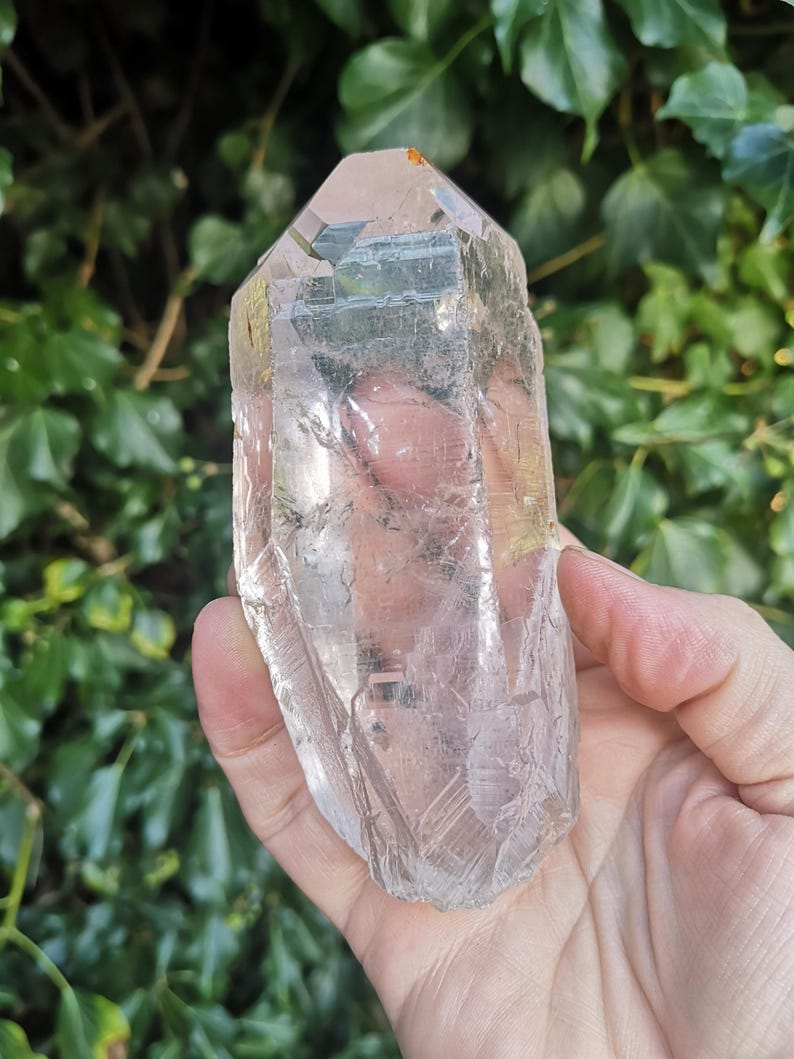 May include: A large, clear quartz crystal with a pointed tip and a smooth, polished surface. The crystal is held in a hand, and the background is blurred.