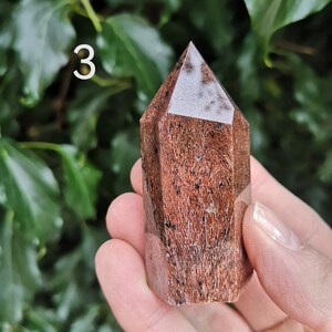 Puede incluir: Una punta de cristal marr&oacute;n y blanco con una parte superior puntiaguda y una base facetada. El cristal mide aproximadamente 5 cm de alto y tiene una textura natural y rugosa.