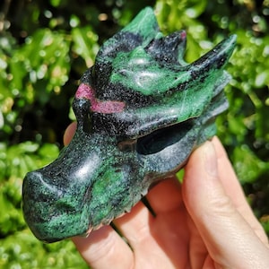 May include: A carved dragon head sculpture in shades of green, black, and red. The dragon's head is detailed with textured scales and features a prominent snout and horns. The sculpture is held in a hand against a blurred green background.