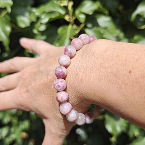 May include: A beaded bracelet featuring smooth, round, polished stones in varying shades of pink, ranging from pale blush to deep magenta, strung together on an elastic cord to form a continuous circular band around the wrist.