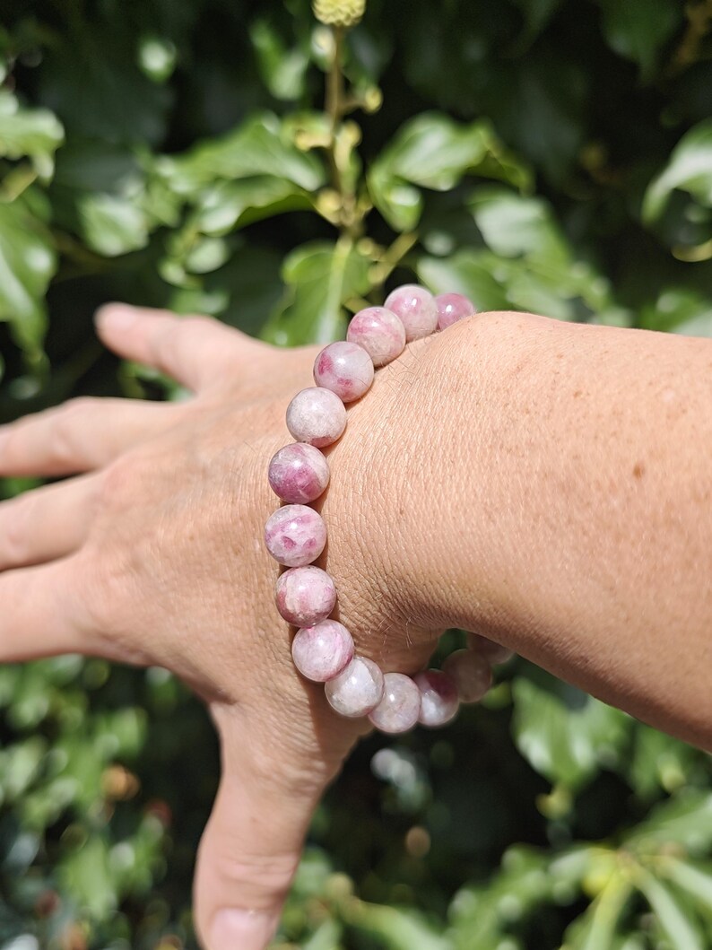 May include: A beaded bracelet featuring smooth, round, polished stones in varying shades of pink, ranging from pale blush to deep magenta, strung together on an elastic cord to form a continuous circular band around the wrist.
