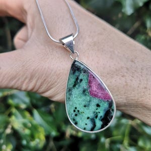 May include: A silver necklace with a teardrop-shaped pendant. The pendant features a green and black stone with a pink section. The necklace is held by a hand against a blurred green background.