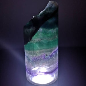 May include: A green, purple, and white fluorite crystal with a light shining through it. The crystal is shaped like a cylinder and has a rough, natural texture.