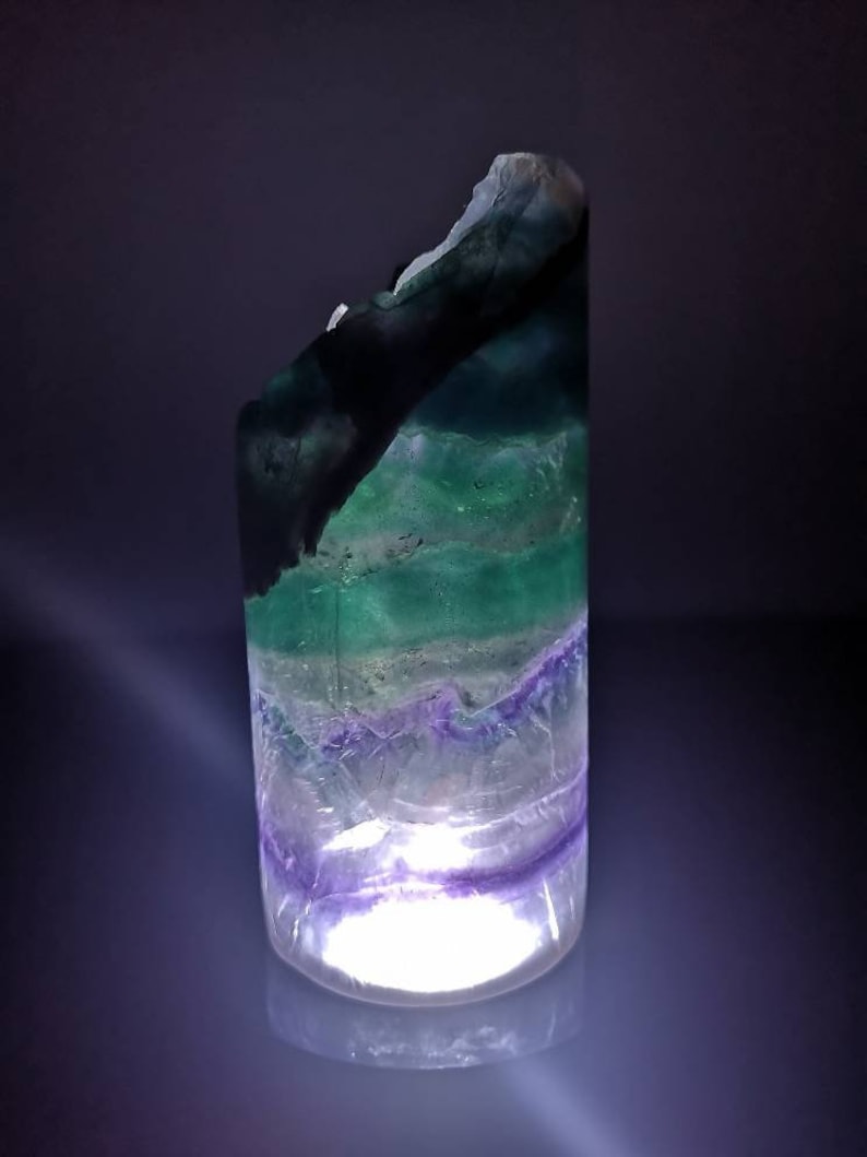May include: A green, purple, and white fluorite crystal with a light shining through it. The crystal is shaped like a cylinder and has a rough, natural texture.