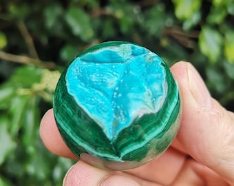 May include: A polished, spherical gemstone featuring a vibrant teal center surrounded by concentric bands of deep green malachite. The stone is held in a hand, showcasing its natural patterns and textures.