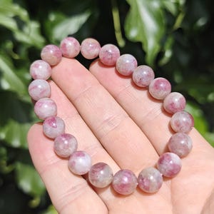 May include: A beaded bracelet featuring round, polished stones in varying shades of pink, white, and translucent grey. The beads are strung together in a circular loop, displaying natural color variations and internal textures within each stone.