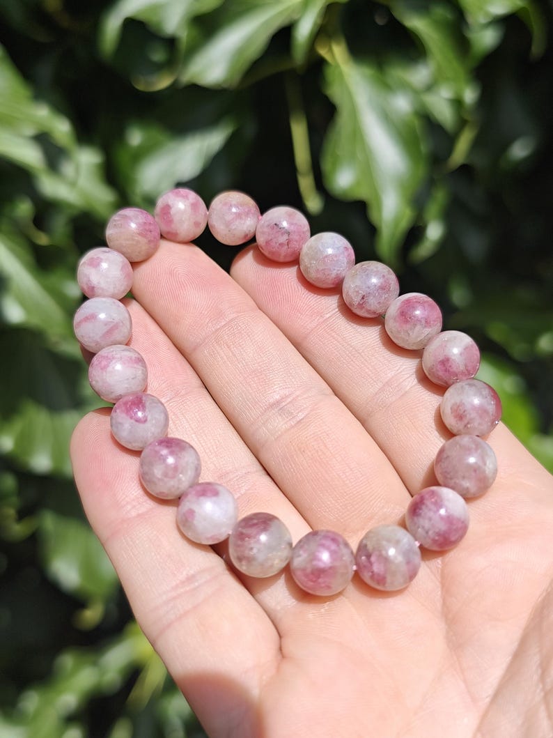 May include: A beaded bracelet featuring round, polished stones in varying shades of pink, white, and translucent grey. The beads are strung together in a circular loop, displaying natural color variations and internal textures within each stone.