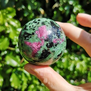May include: A polished sphere of green zoisite with ruby and black tourmaline inclusions. The gemstone ball is held in a hand against a blurred green background. The ruby inclusions form a striking pink pattern.