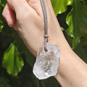 May include: A clear quartz crystal pendant on a gray cord necklace. The irregularly shaped crystal is suspended from a silver-colored metal bead. The background features green foliage.
