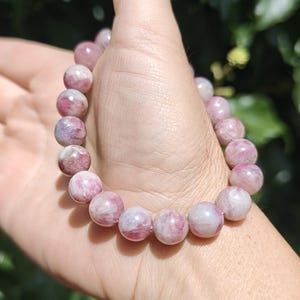 May include: A beaded bracelet featuring smooth, round, polished stones in varying shades of pink, magenta, and white. The beads are strung together in a circular pattern, displaying natural marbling and translucent qualities throughout each stone.