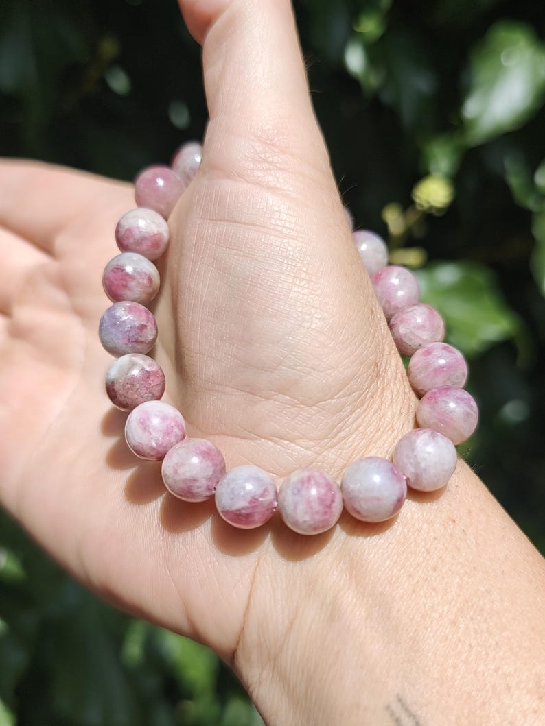 May include: A beaded bracelet featuring smooth, round, polished stones in varying shades of pink, magenta, and white. The beads are strung together in a circular pattern, displaying natural marbling and translucent qualities throughout each stone.