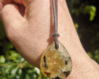May include: A teardrop-shaped pendant necklace with a brown cord. The pendant is a translucent yellow-orange color with black and brown inclusions. A silver-colored bead accents the cord where it meets the pendant.