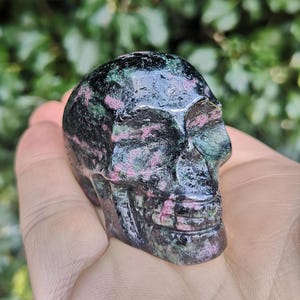 May include: A black, green and pink gemstone skull. The skull is carved from a single piece of stone and has a detailed face.