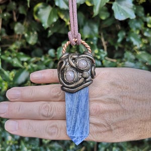 May include: A blue kyanite crystal pendant necklace. The crystal is suspended from a bronze-coloured metal pendant with a decorative design and two round, light-coloured stones. The necklace has a brown cord.