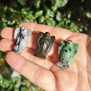 May include: Three carved angel figurines made of different types of stone. The first is a white and black snowflake obsidian angel, the second is a black obsidian angel, and the third is a green aventurine angel.