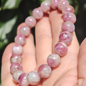 May include: A beaded bracelet composed of round, polished stones in varying shades of pink, magenta, and translucent white. The beads are strung together in a circular formation, displaying natural mineral inclusions and a smooth, glossy surface texture.