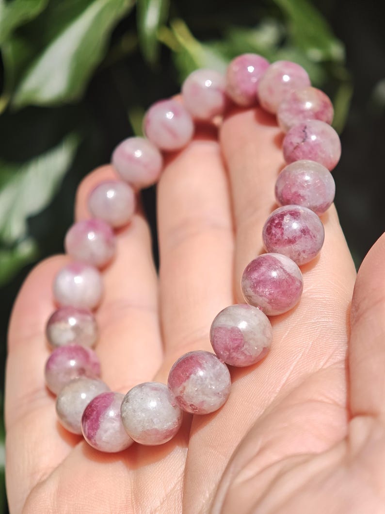 May include: A beaded bracelet composed of round, polished stones in varying shades of pink, magenta, and translucent white. The beads are strung together in a circular formation, displaying natural mineral inclusions and a smooth, glossy surface texture.