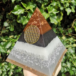 May include: A layered pyramid-shaped orgone device with a gold-colored Sacred Geometry symbol. The pyramid features brown, black, white, and gray layers with metallic accents. The pyramid is held in a hand against a backdrop of green foliage.