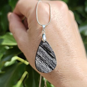 May include: A teardrop-shaped pendant featuring a textured, dark grey and black surface with metallic silver flecks. The pendant is suspended from a thin, silver-coloured snake chain necklace, held by a silver-toned metal bail at the top.