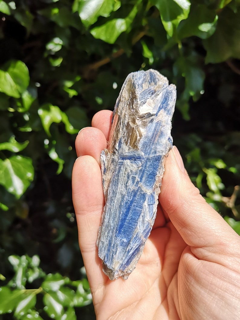 K&ouml;nnte beinhalten: Ein blauer Kyanit-Kristall mit silbernem Schimmer. Der Kristall ist an einem Ende spitz und hat eine raue, nat&uuml;rliche Textur.