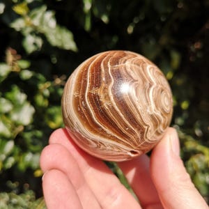 K&ouml;nnte beinhalten: Eine glatte, polierte braune und wei&szlig;e, geb&auml;nderte Kugel aus geb&auml;ndertem Achat. Die Kugel ist etwa so gro&szlig; wie ein Golfball und hat ein nat&uuml;rliches Loch, das durch sie gebohrt wurde.