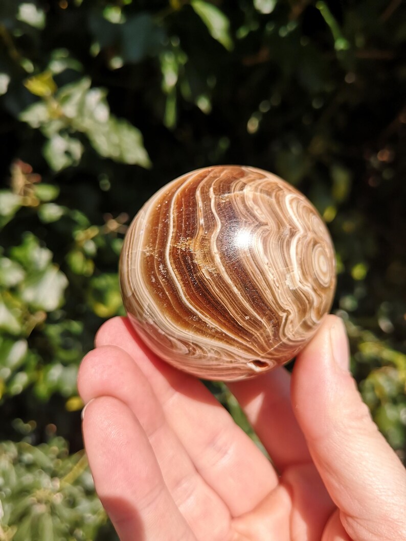 K&ouml;nnte beinhalten: Eine glatte, polierte braune und wei&szlig;e, geb&auml;nderte Kugel aus geb&auml;ndertem Achat. Die Kugel ist etwa so gro&szlig; wie ein Golfball und hat ein nat&uuml;rliches Loch, das durch sie gebohrt wurde.