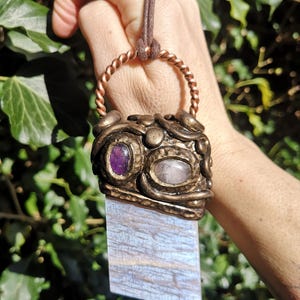 May include: A handcrafted pendant necklace with a rectangular, iridescent blue-grey stone. The pendant features bronze-coloured metalwork, two oval gemstones, and a twisted copper ring. The necklace has a brown cord.