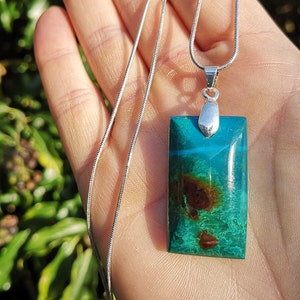 Pu&ograve; includere: Una collana con pendente rettangolare con una pietra turchese e marrone. La pietra &egrave; incastonata in metallo color argento e pende da una delicata catena d'argento. Il pendente ha una finitura lucida e brillante.