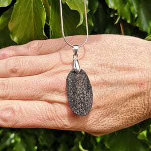 May include: A silver necklace featuring a dark grey, oval pendant with a textured surface. The pendant hangs from a fine silver chain. Green foliage forms the backdrop.