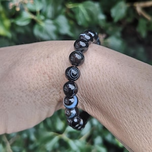 May include: A black and white beaded bracelet. The bracelet features round beads with a glossy finish. The beads have a marbled appearance with concentric circles and swirls. The bracelet is worn on a wrist.